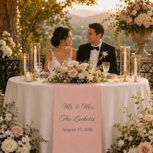 以下が含まれることがあります： 薄いピンク色のテーブルランナー。黒い文字で「Mr. & Mrs. The Lucketts August 15, 2026」とプリントされており、白いテーブルクロスをかけた円卓に掛けられています。花のアレンジメント、ガラス製のキャンドルホルダー、白いキャンドルで飾られています。
