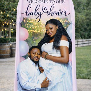 May include: A baby shower banner with a pink arch design. The banner features a couple, with the pregnant woman in a white dress and the man in a blue shirt. The text "WELCOME TO OUR baby shower" is displayed in elegant script.
