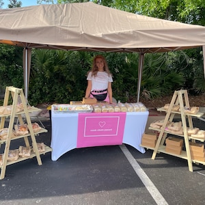 May include: A woman stands behind a table at an outdoor market stall. The table is covered with a white cloth and a pink banner that reads "cook clean parent." Baked goods are displayed on the table and on wooden tiered shelves.