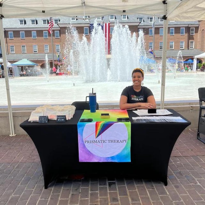 May include: A person sits behind a table with a black tablecloth and a rainbow banner that reads "Prismatic Therapy". The table is set up in front of a fountain with a brick building in the background.