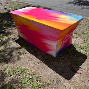 May include: A rectangular fitted table cover featuring a vibrant, multi-colored abstract print with shades of pink, orange, yellow, and blue. The fabric displays the text J+M Prints and a QR code on the top surface, creating a smooth, taut appearance.