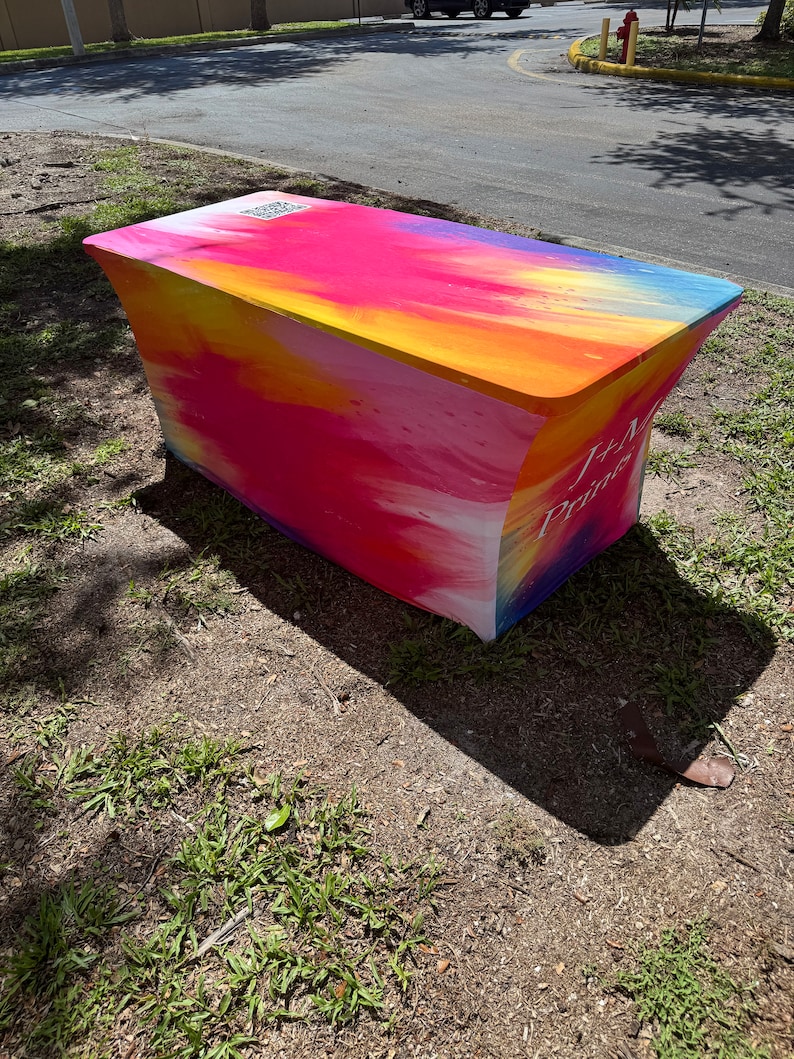 May include: A rectangular fitted table cover featuring a vibrant, multi-colored abstract print with shades of pink, orange, yellow, and blue. The fabric displays the text J+M Prints and a QR code on the top surface, creating a smooth, taut appearance.