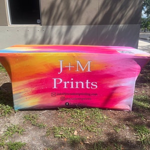 May include: A rectangular fitted table cover featuring a vibrant watercolor-style gradient of pink, orange, yellow, and blue. The text J+M Prints is centered in white, accompanied by an email address, Instagram handle, and Facebook page name.