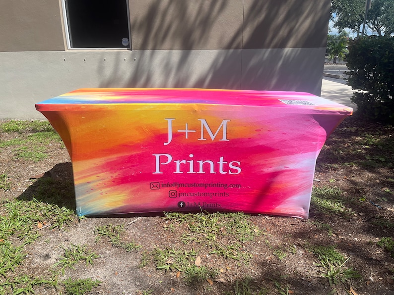 May include: A rectangular fitted table cover featuring a vibrant watercolor-style gradient of pink, orange, yellow, and blue. The text J+M Prints is centered in white, accompanied by an email address, Instagram handle, and Facebook page name.