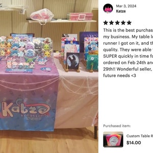 May include: A pink and blue table runner with the text "Katze Kreations" printed on it. The table runner is decorated with a white pearl garland. The table runner is displayed on a table with various items for sale, including a clear acrylic display case filled with colorful character figurines, a framed painting, and a sign with the text "Katze Kreations".