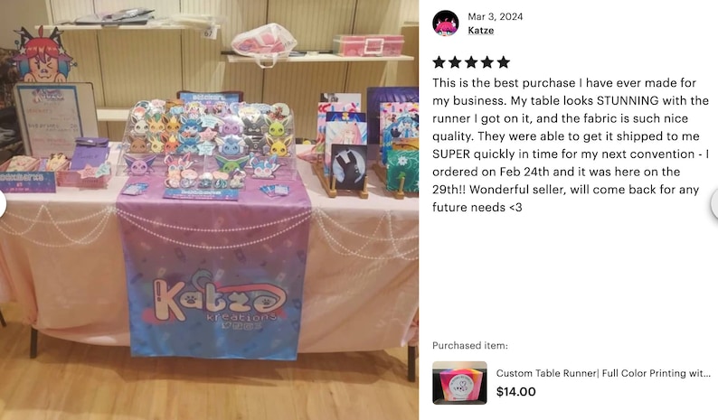 May include: A pink and blue table runner with the text "Katze Kreations" printed on it. The table runner is decorated with a white pearl garland. The table runner is displayed on a table with various items for sale, including a clear acrylic display case filled with colorful character figurines, a framed painting, and a sign with the text "Katze Kreations".