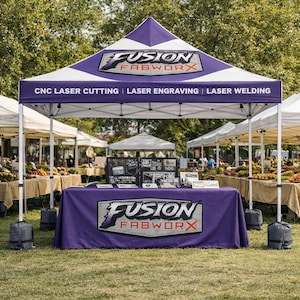 May include: A purple and white canopy tent displaying the "Fusion Fabworx" logo and services: CNC laser cutting, laser engraving, and laser welding. The tent is at an outdoor market, with other stalls and tables visible.