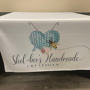 May include: White table runner with a blue and white knitted heart design, a bee, and the text "Shel-bee's Handmade Creations".