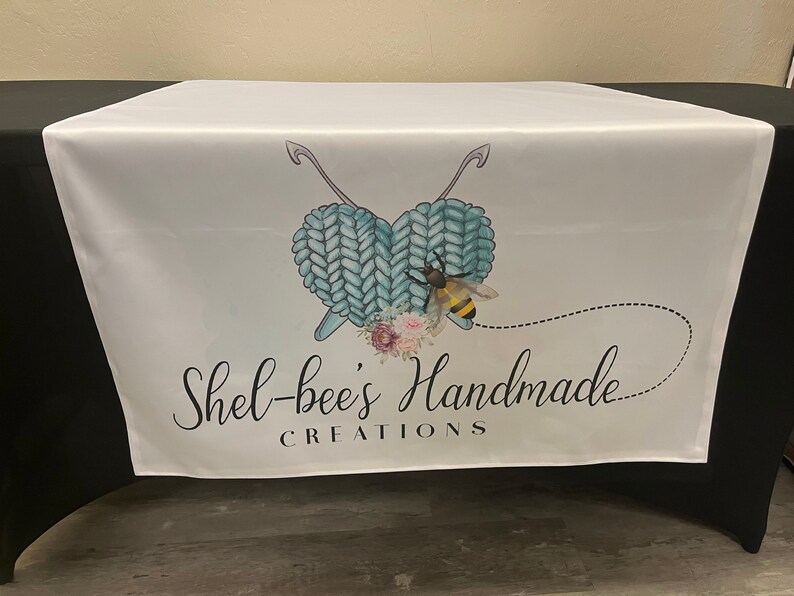 May include: White table runner with a blue and white knitted heart design, a bee, and the text "Shel-bee's Handmade Creations".
