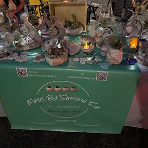 May include: A table display of handmade concrete, resin, and wood items. The items include planters, ornaments, and candle holders. The table has a green tablecloth with the text "Made By Sammy Sue" and contact information.