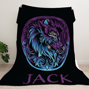 Op de afbeelding: Zwarte fleece deken met een illustratie van een paarse en blauwe wolf in een cirkelvormig frame met sierlijke details. De naam "JACK" is in paars onder de wolf gedrukt.