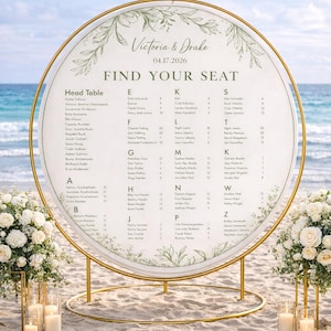 Op de afbeelding: Een gouden, cirkelvormige zitplaatsindeling voor een bruiloft op het strand. Het bord toont namen alfabetisch, met de trouwdatum 17.04.2026 en de namen Victoria & Drake bovenaan. Bloemschikkingen en kaarsen flankeren het bord.