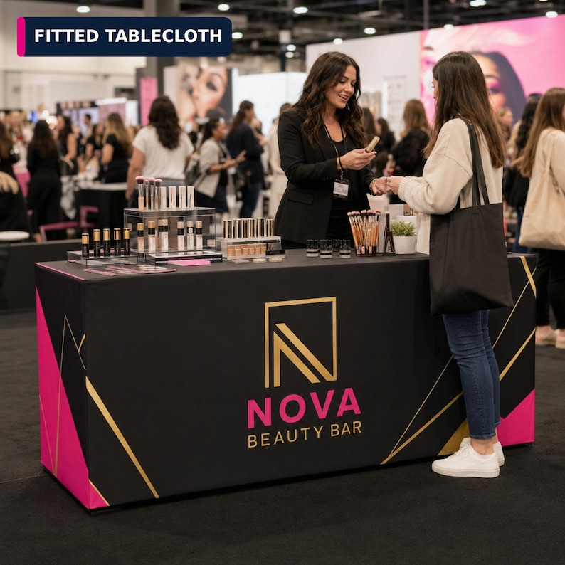 May include: A black fitted tablecloth featuring a gold and pink logo that reads NOVA BEAUTY BAR. The fabric cover has a geometric design with gold lines and pink triangular accents on the corners, designed to fit a rectangular display table.