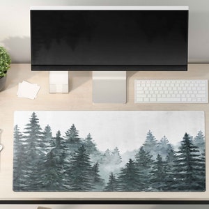 Puede incluir: Una alfombrilla de escritorio blanca con un diseño de acuarela de un bosque brumoso con árboles verde oscuro. La alfombrilla está sobre un escritorio de madera clara con un monitor de ordenador, un teclado, un ratón, una planta, un reloj y un vaso de agua.