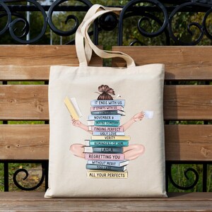 May include: A cream-coloured tote bag with a graphic of a person sitting on a pile of books. The books are titled "It Ends With Us" and "November 9." The person holds a book and a cup. The bag has long handles.