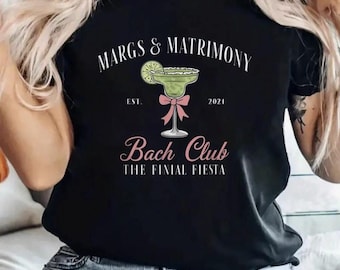 Puede incluir: Camiseta negra con un gráfico blanco de un vaso de margarita con una rodaja de lima y un lazo rosa. El texto "Margs & Matrimony Est. 2024 Bach Club The Final Fiesta" está impreso en la camiseta.