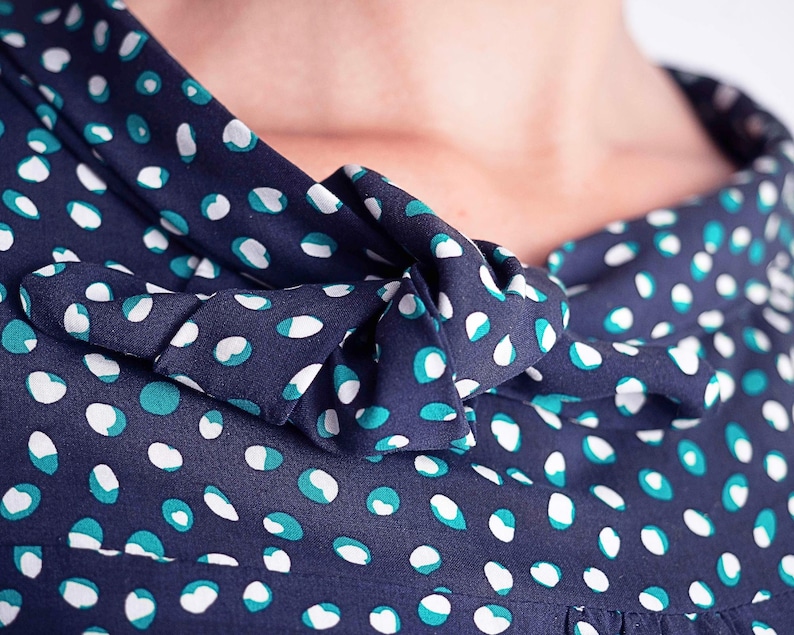 May include: Close-up of a navy blue fabric with a white and teal polka dot pattern. The fabric is gathered at the neckline and forms a bow. The image focuses on the details of the fabric and its design.