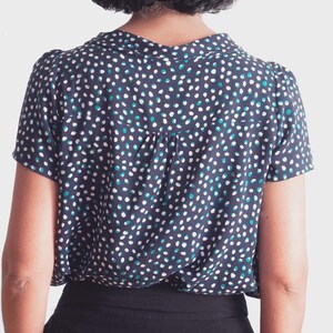 May include: A navy blue short-sleeved blouse with a white, teal, and blue polka dot pattern. The blouse has a collared neckline and a pleated back detail. The bottom portion of the blouse is black.