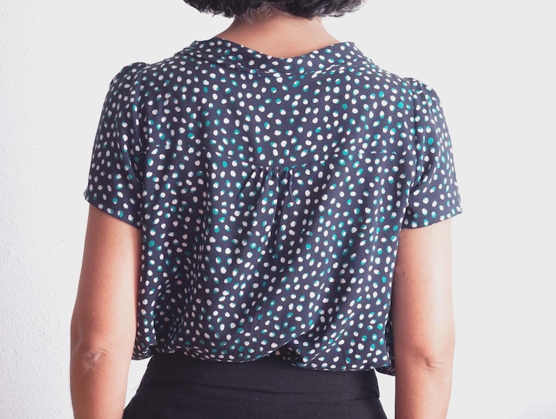 May include: A navy blue short-sleeved blouse with a white, teal, and blue polka dot pattern. The blouse has a collared neckline and a pleated back detail. The bottom portion of the blouse is black.