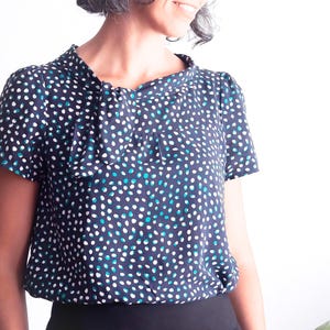 May include: A navy blue blouse with a pattern of white and teal dots. The short-sleeved top has a gathered neckline and a slightly puffed sleeve. The bottom of the blouse is gathered at the waist.