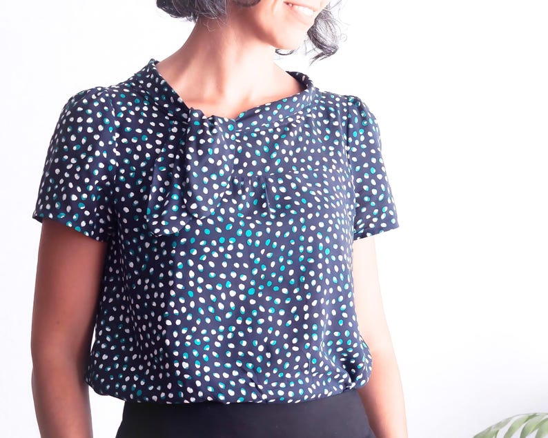 May include: A navy blue blouse with a pattern of white and teal dots. The short-sleeved top has a gathered neckline and a slightly puffed sleeve. The bottom of the blouse is gathered at the waist.