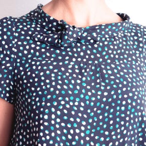 May include: Navy blue short-sleeved top with a polka dot pattern in white and teal. The top features a high, ruffled collar with a bow detail. The fabric appears to be lightweight and suitable for warmer weather.