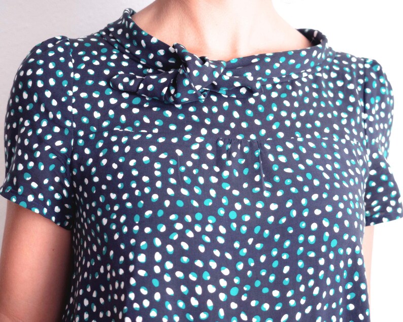 May include: Navy blue short-sleeved top with a polka dot pattern in white and teal. The top features a high, ruffled collar with a bow detail. The fabric appears to be lightweight and suitable for warmer weather.