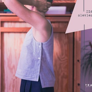 Op de afbeelding: Een lichtgrijs mouwloos topje met drie witte knopen aan de zijkant. De top wordt gecombineerd met een donkerblauwe rok. De afbeelding bevat de tekst "IDA sleeveless top" en het logo "TRAMA patternstudio".