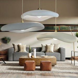 May include: Three pendant lights with irregular, organic, cloud-like shapes suspended from the ceiling. Each fixture features a smooth, matte light grey finish, a central light bulb, and a thin metallic cord connecting to a brass-toned ceiling mount.