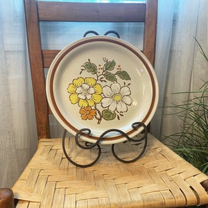 May include: A vintage floral dinner plate with a brown rim and a white center featuring yellow, orange, and white flowers. The plate is on a wooden chair with a woven seat.