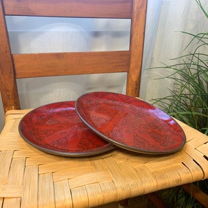 May include: Two red ceramic dinner plates with a black floral design. The plates are stacked on top of each other and are sitting on a woven chair seat.