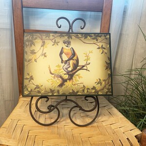 May include: A framed illustration of a monkey sitting on a branch with green leaves. The frame is on a black metal easel.