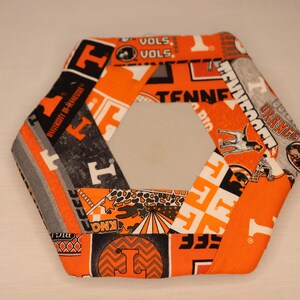 May include: A hexagonal fabric coaster featuring orange, gray, and black patterns. The design includes the text "VOLS," "TENNESSEE," and "UNIVERSITY OF TENNESSEE." The coaster is a decorative item.