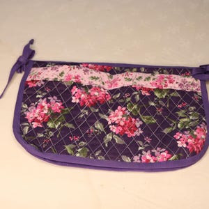 May include: A quilted, floral-patterned apron in shades of purple, pink, and green. The apron features a dark purple base with a diamond stitch pattern and a floral design. It has a purple border and ties.