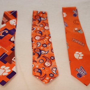 May include: Three orange neckties with different patterns. One tie has a geometric design with a paw print and the letter "C". Another has a paisley pattern. The third tie has the word "Tigers" and a basketball graphic.