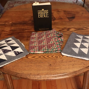 May include: A wooden table displays a black Bible with gold lettering, flanked by two square quilted trivets with triangle patterns in black, white, and paisley prints. A folded plaid fabric in red, green, and white is between them.