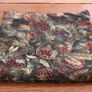 May include: A folded square of fabric with a dark background featuring a pattern of pinecones, pine needles, and leaves in shades of brown, green, and cream. The fabric is likely cotton or a similar material.