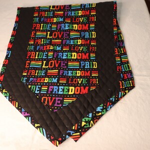 May include: A quilted table runner with a black border and a central panel featuring the words "PRIDE", "FREEDOM", and "LOVE" in rainbow colors on a black background. The runner is made of fabric.