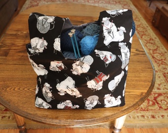 May include: A black tote bag with a sheep pattern, filled with blue and gray yarn, and knitting needles. The bag is sitting on a wooden table. The sheep are white with various colored sweaters and holding knitting needles.