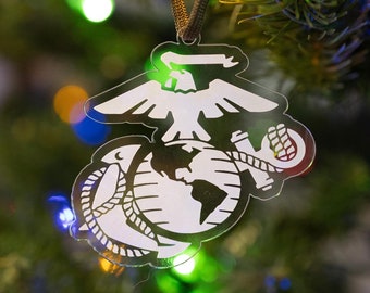 May include: A clear, decorative Christmas ornament featuring the emblem of the United States Marine Corps. The ornament is white with black outlines and hangs from a brown cord. The background is a Christmas tree with colorful lights.