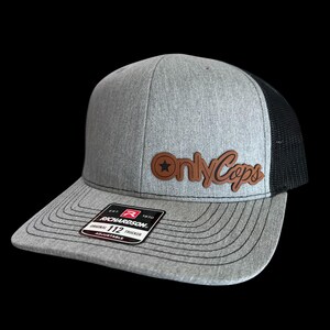 May include: A gray Richardson 112 trucker hat with a black mesh back. The hat features a brown leather patch with the words "Only Cops" and a star. The brim is gray with a curved design.