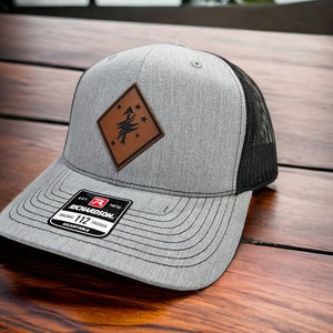 May include: A gray and black trucker hat with a brown leather patch featuring a stylized tree and stars. The patch has the text "112" on it. The hat has a black mesh back and an adjustable strap with the text "Richardson 112 Trucker Adjustable" on it.