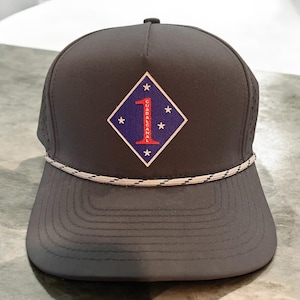 May include: A dark gray baseball cap with a flat brim and a white and gray rope detail. The cap features a diamond-shaped patch with a red number 1, the word GUADALCANAL, and stars on a blue background.