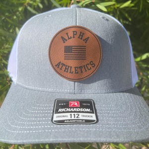 May include: A gray and white trucker hat with a brown leather patch that says "ALPHA ATHLETICS" with a small American flag inside the patch. The hat also has a black and red label that says "RICHARDSON" with the text "EST. 1970 ORIGINAL 112 TRUCKER ADJUSTABLE".