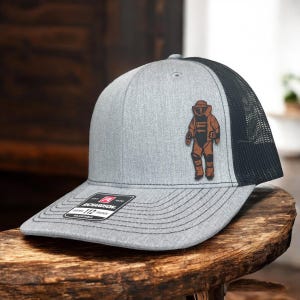 May include: A gray and black trucker hat with a brown patch of a person in a bomb suit. The hat has a curved brim and a mesh back. The patch is on the front of the hat. The hat has a tag that says "Richardson 112".