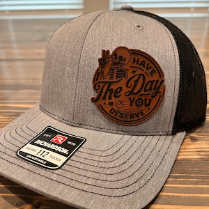 May include: Gray and black trucker hat with a brown leather patch that says "Have The Day You Deserve" with a skeleton holding a peace sign. The patch is stitched onto the hat. The hat has a Richardson 112 label on the side.