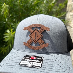 May include: A gray and black trucker hat with a brown leather patch featuring the text "1st Battalion 11th Marines", a crossed cannon design, and the number "1". The patch also includes the text "Ultima Ratio Regum". The hat has a black mesh back and a Richardson 112 adjustable strap.