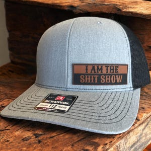 May include: Gray trucker hat with a black mesh back and a curved brim. The hat features a brown and black patch that reads "I AM THE SHIT SHOW". A Richardson 112 Trucker tag is visible.