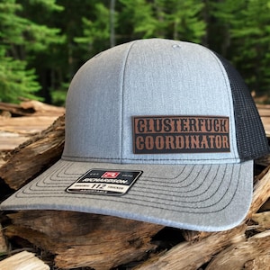 May include: Gray trucker hat with a black mesh back and a curved brim. The hat features a brown leather patch with the words "CLUSTERFUCK COORDINATOR" in a bold, Western-style font. The hat is adjustable and has a Richardson 112 tag.