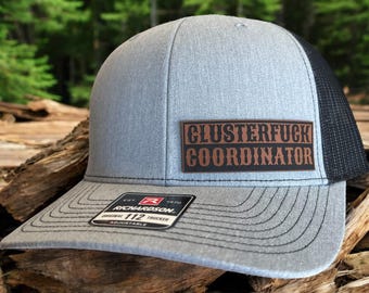 Puede incluir: Gorra de camionero gris con parte trasera de malla negra y visera curva. La gorra presenta un parche de cuero marrón con las palabras "CLUSTERFUCK COORDINATOR" en una fuente audaz de estilo occidental. La gorra es ajustable y tiene una etiqueta Richardson 112.